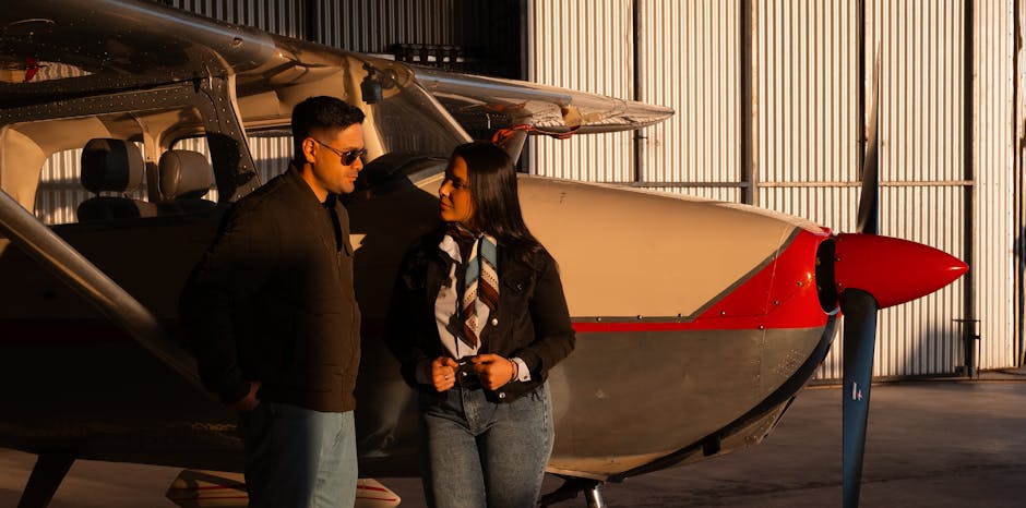Pilot sitting in an aircraft cockpit inside a hangar with a crew member in the background.